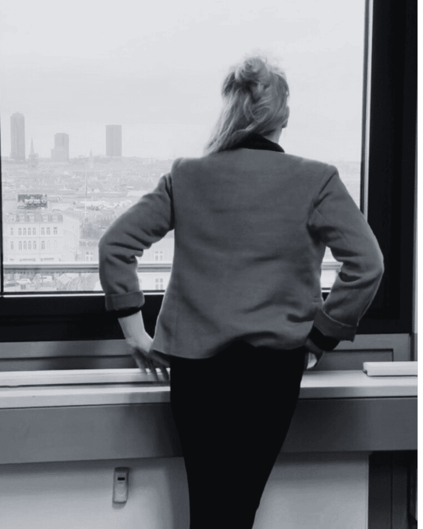 Person with long hair in a jacket standing indoors, looking out a window at a cityscape with tall buildings in the background.