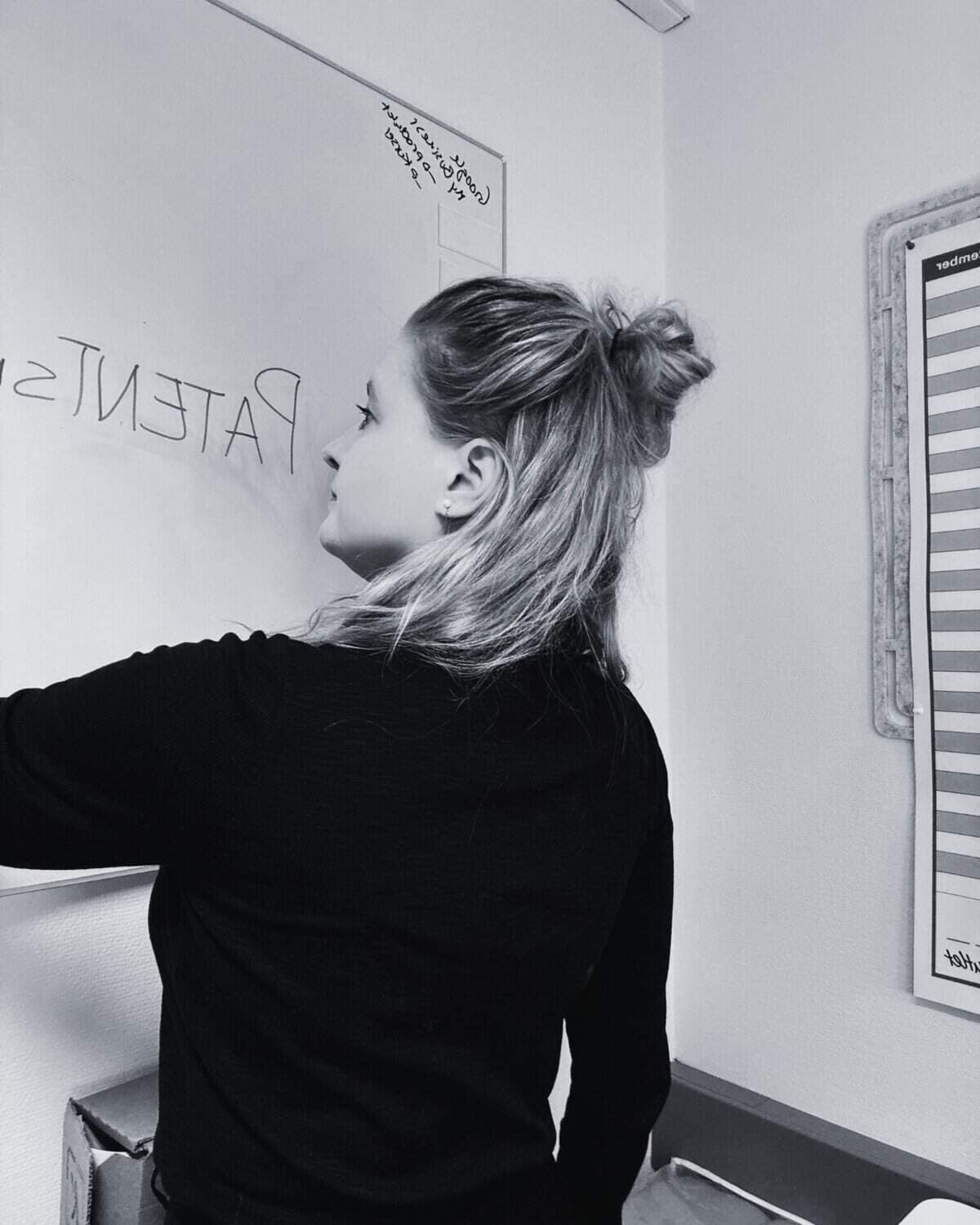 A person with long hair in a bun writes on a whiteboard in a room, partially visible calendar chart on the wall.