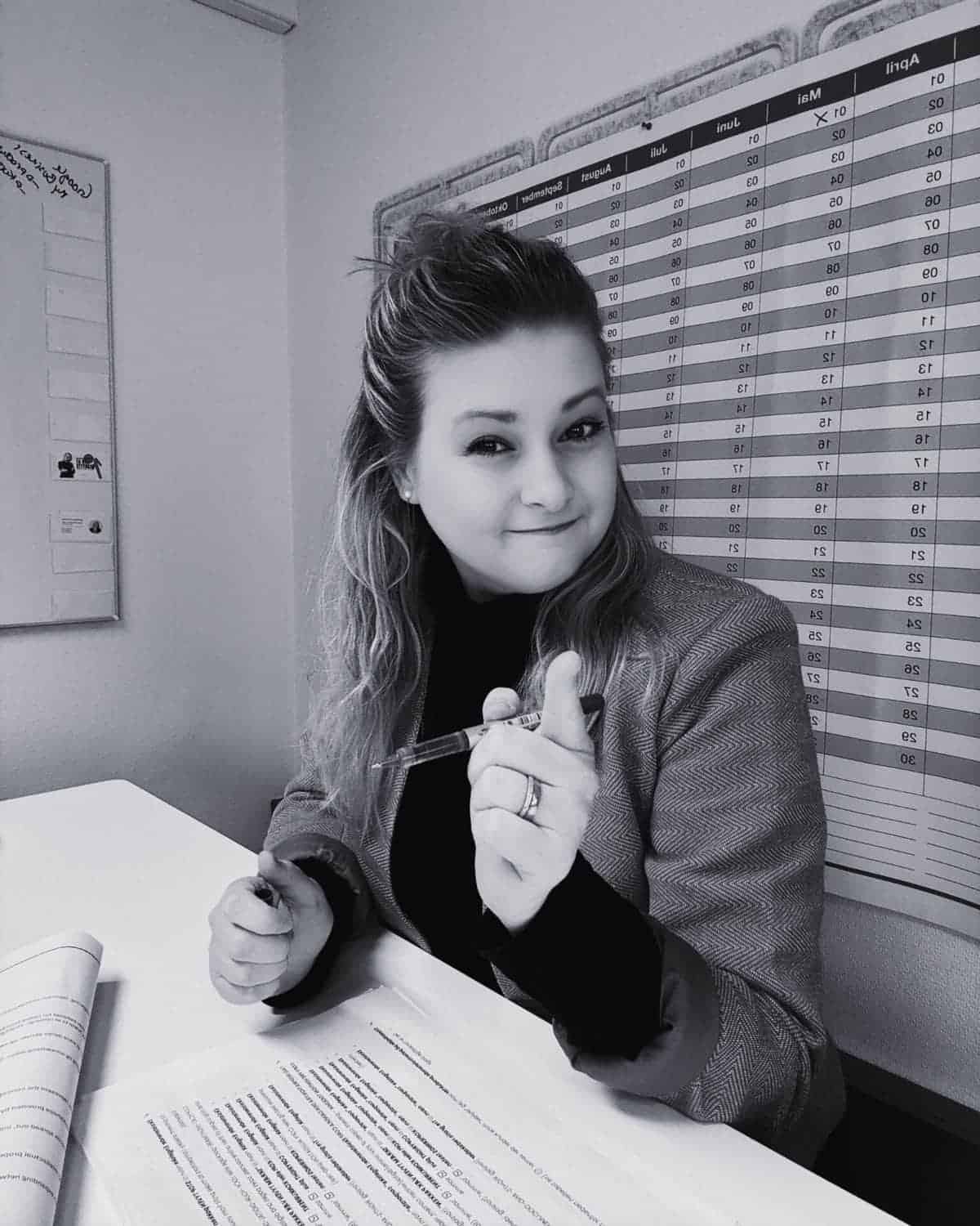 Gyde balzer carstensen, your intellectual property rights mentor and coach is sitting at a desk, holding a pen and pointing, with a large calendar on the wall behind them. Papers are spread out on the desk.