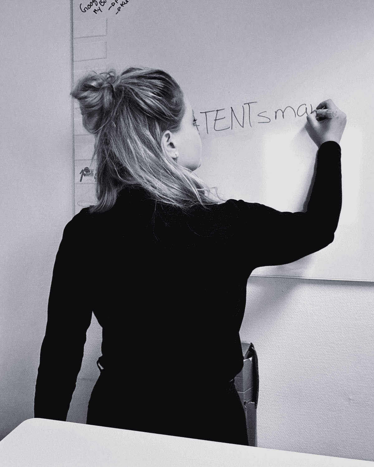 Person with long hair in a ponytail, wearing a dark top, writes on a whiteboard with a marker in a classroom or office setting. The photo is in black and white.