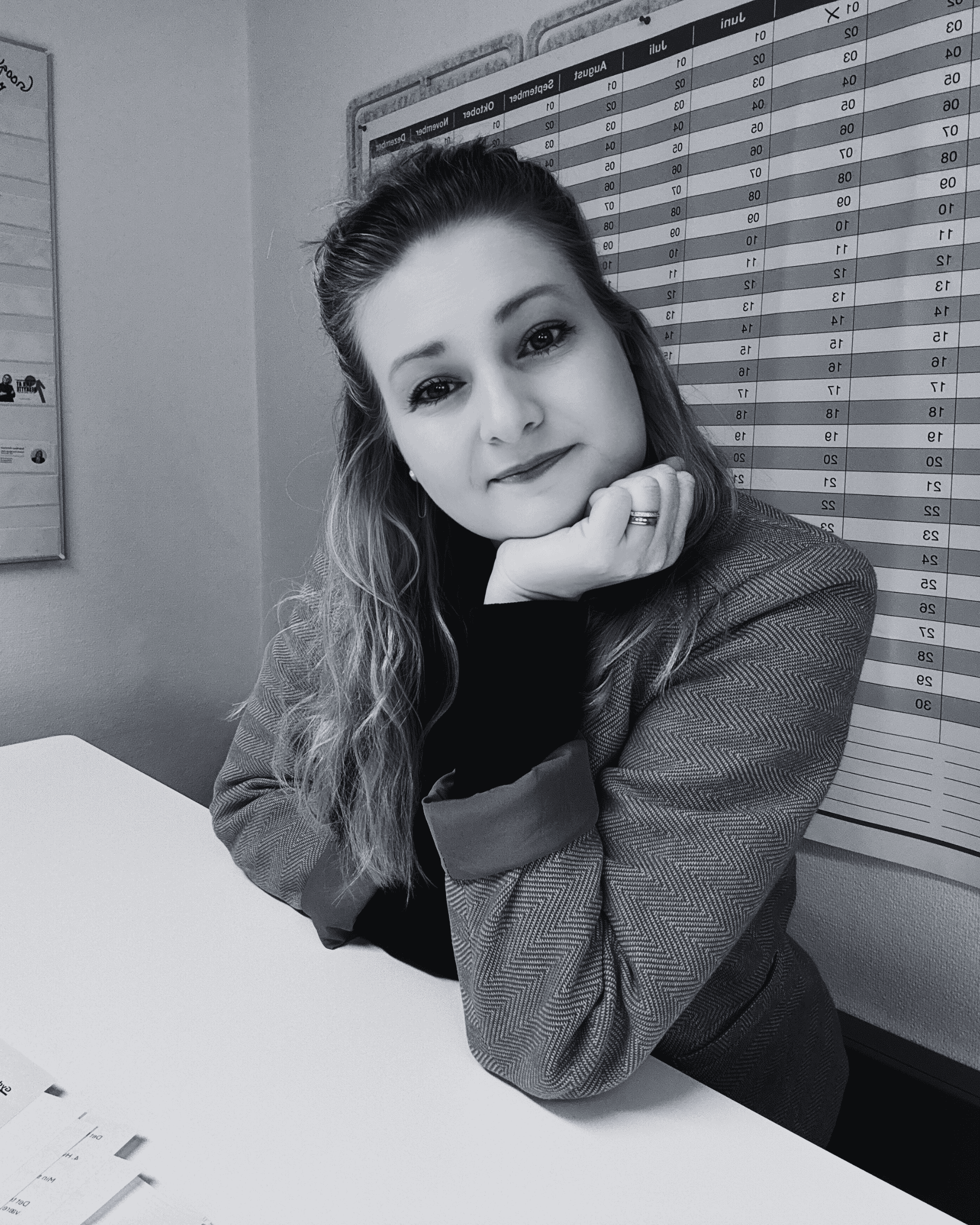 Gyde Balzer Carstensen in a blazer sits at her desk, resting her chin on her hand, with a calendar marking key dates for IP strategy in the background. Her gaze meets the camera in this captivating black and white image.